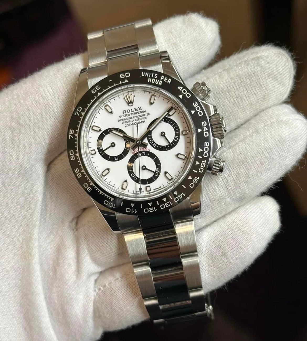 Person wearing white gloves holding a Rolex Daytona watch with a silver bracelet and white dial.